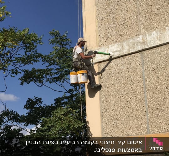 אדם צובע קיר חיצוני בעזרת רולר על חבלים